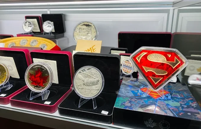 A shop display case: a Canada 2019 red maple leaf coin, a large silver coin, boxed silver rounds and a Superman shield coin.